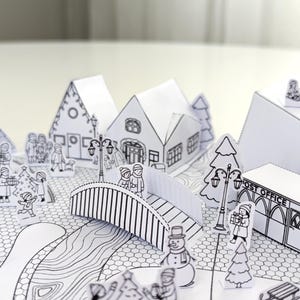 May include: A black and white papercraft winter village scene. The scene includes houses, a bridge, trees, and figures of people. The post office is visible. The overall theme is a winter holiday scene.