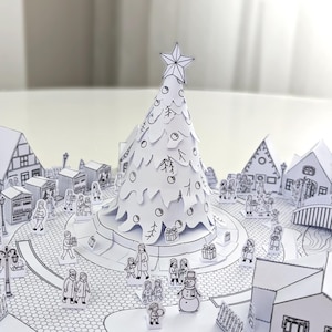 May include: A detailed papercraft Christmas scene featuring a central tree topped with a star. The white tree is surrounded by a town with houses, shops, and people. The scene is entirely in black and white, creating a charming, handcrafted aesthetic.