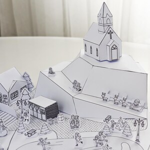 May include: A detailed, black and white paper diorama of a winter village scene. The scene includes a church, houses, trees, and figures engaged in winter activities like skiing and ice skating. Santa Claus and reindeer are also present.