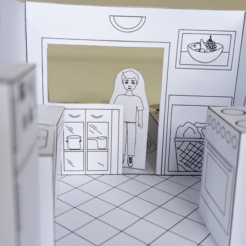 May include: A paper craft kitchen playset featuring black and white line art. The scene includes a flat cutout figure of a person, a stove, cabinets with pots, a laundry basket, and a framed bowl of fruit on the wall, all set on a tiled floor.