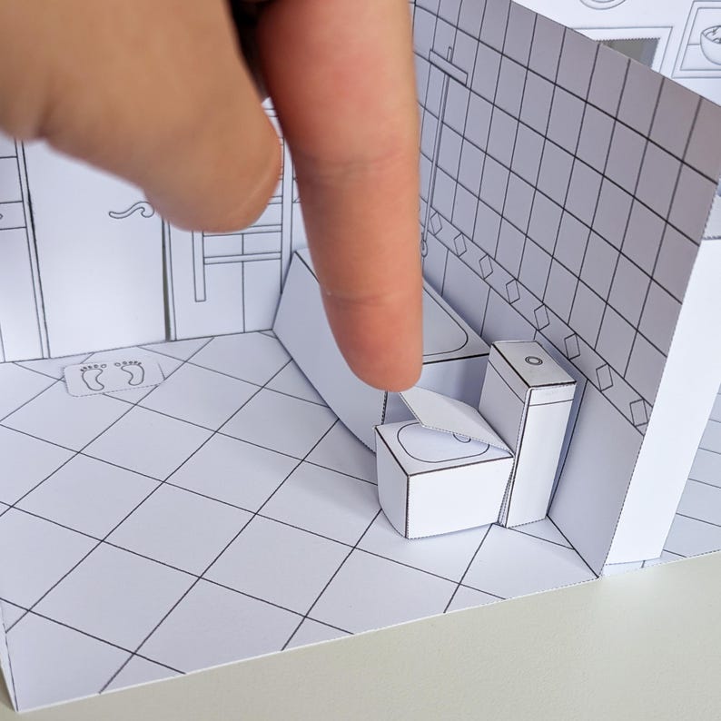 May include: A miniature paper craft bathroom set featuring a white toilet with a movable lid, a bathtub, and tiled floor patterns. The structure is constructed from white cardstock with black line drawings detailing the bathroom fixtures.