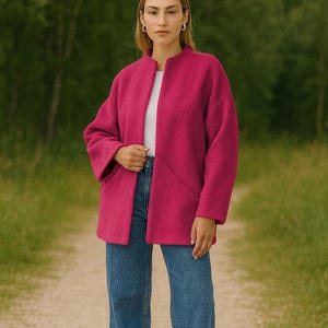 May include: A woman wearing a fuchsia-colored coat over a white top and blue jeans. The coat has a collarless design and long sleeves. The woman is standing on a dirt path with a green background.