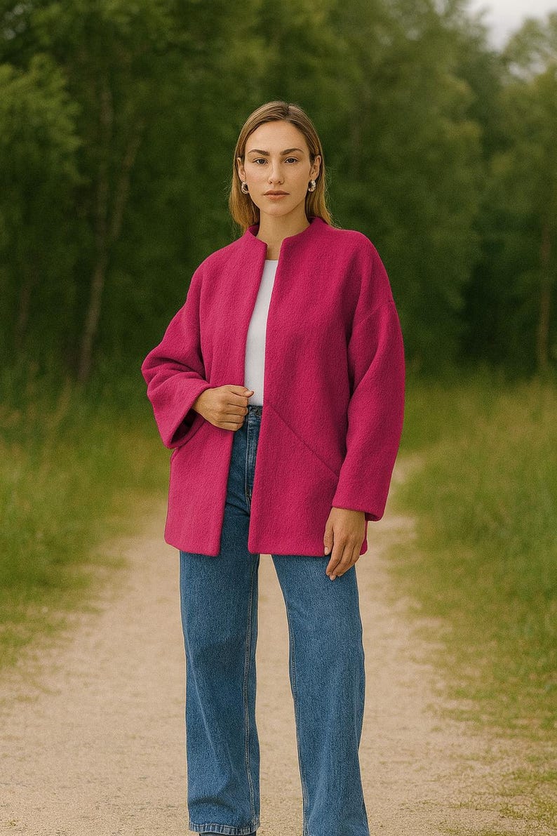 May include: A woman wearing a fuchsia-colored coat over a white top and blue jeans. The coat has a collarless design and long sleeves. The woman is standing on a dirt path with a green background.