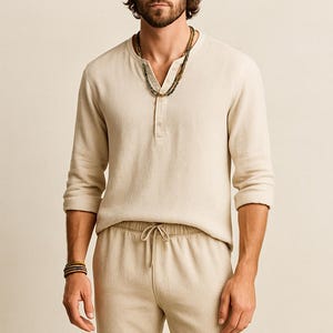 May include: A man wearing a beige linen Henley shirt with three buttons, matching drawstring pants, and a straw hat. He is also wearing a beaded necklace and a bracelet. The outfit is a neutral color.