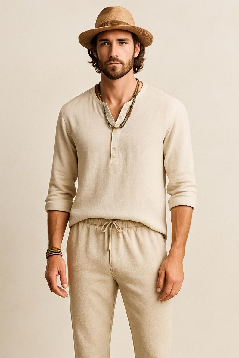 May include: A man wearing a beige linen Henley shirt with three buttons, matching drawstring pants, and a straw hat. He is also wearing a beaded necklace and a bracelet. The outfit is a neutral color.