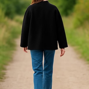 May include: A black jacket with a high collar and long sleeves, paired with blue jeans. The person is walking away on a dirt path, with a blurred green background of trees and foliage.