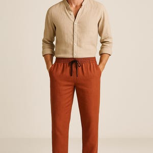 May include: A beige linen shirt with a mandarin collar and long sleeves, paired with rust-colored drawstring pants. The pants have a black drawstring. The outfit is completed with brown sandals.