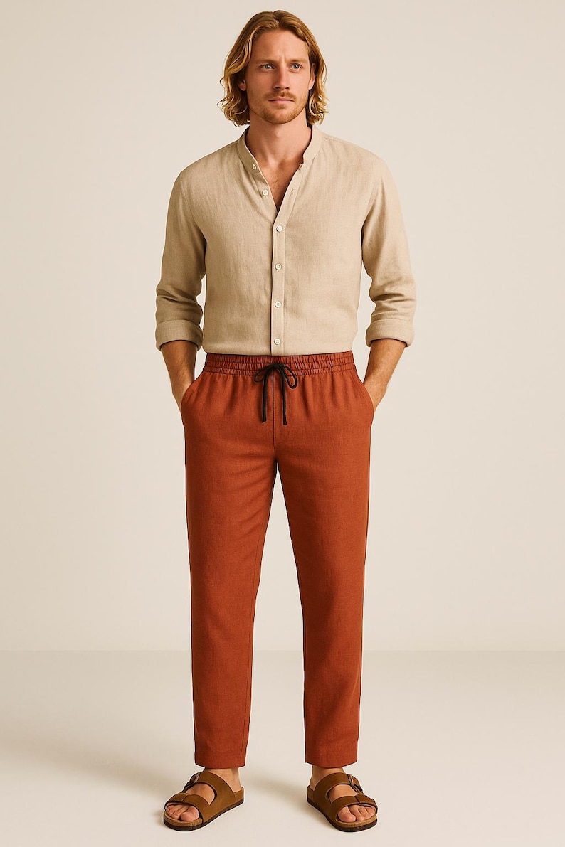 May include: A beige linen shirt with a mandarin collar and long sleeves, paired with rust-colored drawstring pants. The pants have a black drawstring. The outfit is completed with brown sandals.