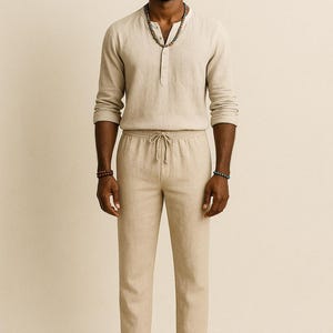 May include: A light beige linen outfit consisting of a long-sleeve Henley shirt, drawstring pants, and a straw fedora hat. The shirt has a single chest pocket. The pants have a relaxed fit. The outfit is suitable for warm weather.