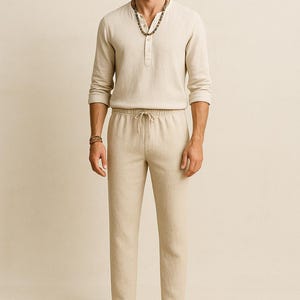 May include: A beige linen outfit consisting of a long-sleeved Henley shirt and drawstring pants. The model is wearing a brown fedora hat and a beaded necklace. The outfit is casual and suitable for warm weather.