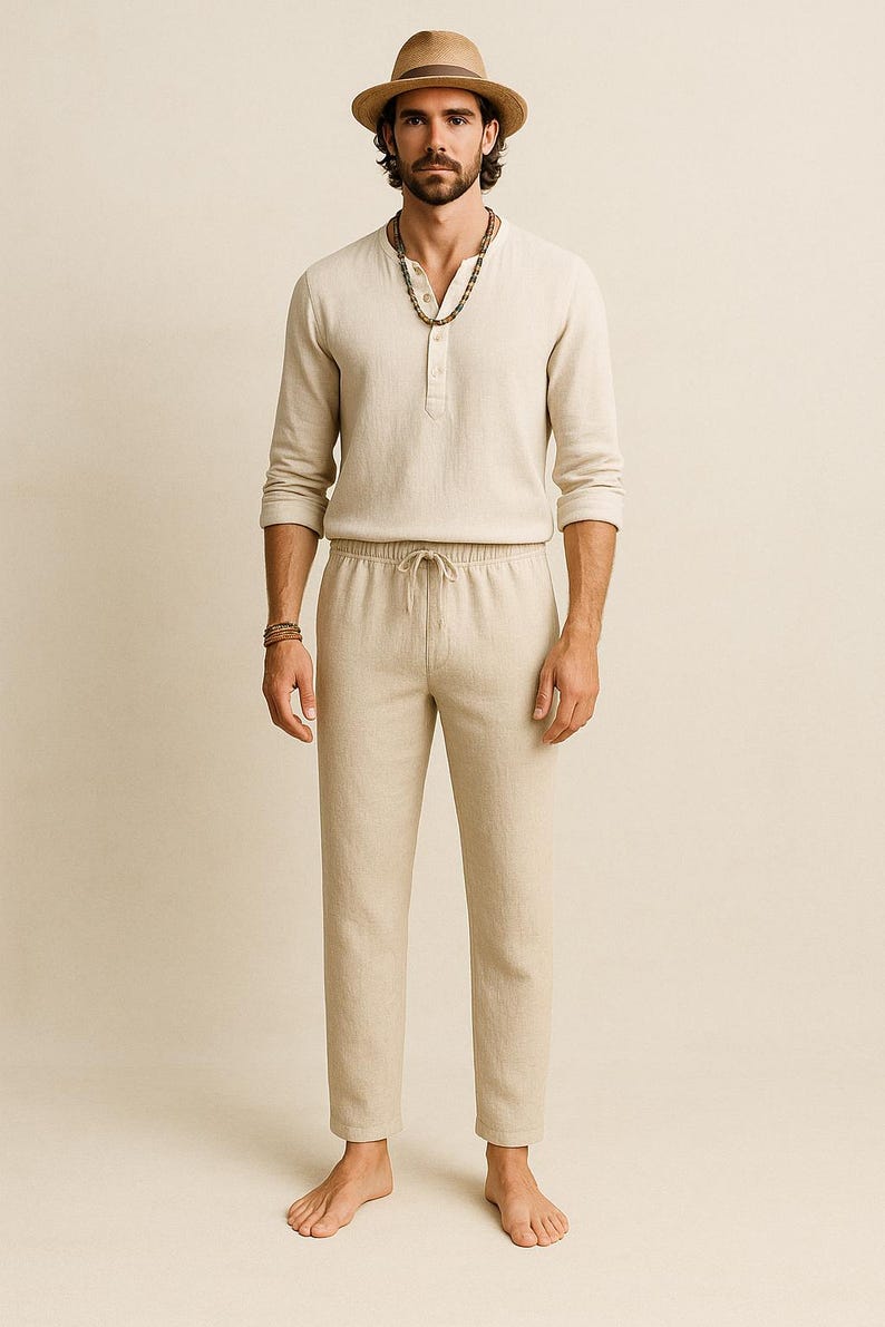 May include: A beige linen outfit consisting of a long-sleeved Henley shirt and drawstring pants. The model is wearing a brown fedora hat and a beaded necklace. The outfit is casual and suitable for warm weather.