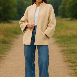 May include: A woman wearing a beige jacket with a mandarin collar, a white top, and blue jeans. The jacket has large pockets and a relaxed fit. The background is a path through a field with trees.
