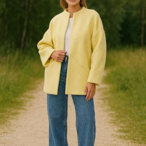 May include: A woman wearing a pale yellow jacket and blue jeans. The jacket has a round collar and long sleeves. The jeans are a medium wash with a wide leg. The background is a blurred green forest.