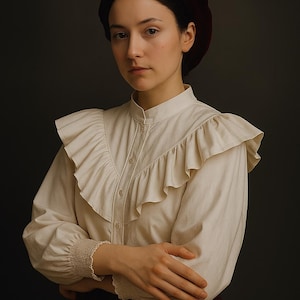May include: A woman wearing a cream-colored blouse with ruffled detailing and a burgundy beret. The blouse has long sleeves with a gathered cuff. The woman is looking directly at the camera.