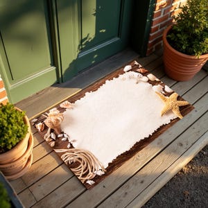 Könnte beinhalten: Eine braune Fußmatte mit einem weißen, rechteckigen Stoffstück in der Mitte. Der Stoff ist mit Muscheln und einem Seestern verziert. Die Fußmatte liegt auf einem Holzboden.