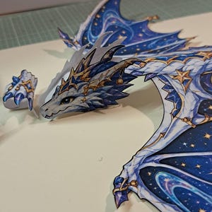 May include: A flat, paper-like cutout of a dragon featuring a white and blue color palette with gold star accents. The dragon has detailed wings with a dark blue night sky pattern, gold star motifs, and white edges. A separate claw piece is included.