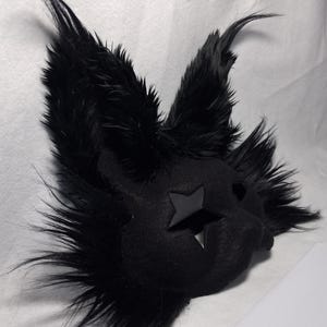 May include: A black, furry mask with pointed ears and a star-shaped cutout on the face. The mask has a plush texture and is likely designed for costume or cosplay purposes. The ears and face are covered in long, black fur.