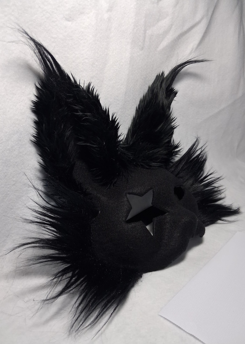 May include: A black, furry mask with pointed ears and a star-shaped cutout on the face. The mask has a plush texture and is likely designed for costume or cosplay purposes. The ears and face are covered in long, black fur.