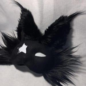 May include: Black felt mask with a star cutout and white eye openings. The mask is adorned with long, black faux fur, giving it a dramatic, animalistic appearance. This costume accessory is suitable for cosplay or themed events.
