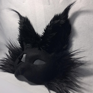 May include: A black, furry mask with pointed ears and eye cutouts. The mask has a smooth, matte finish and is surrounded by long, textured fur. The ears are upright and prominent, giving the mask a stylized animal appearance.