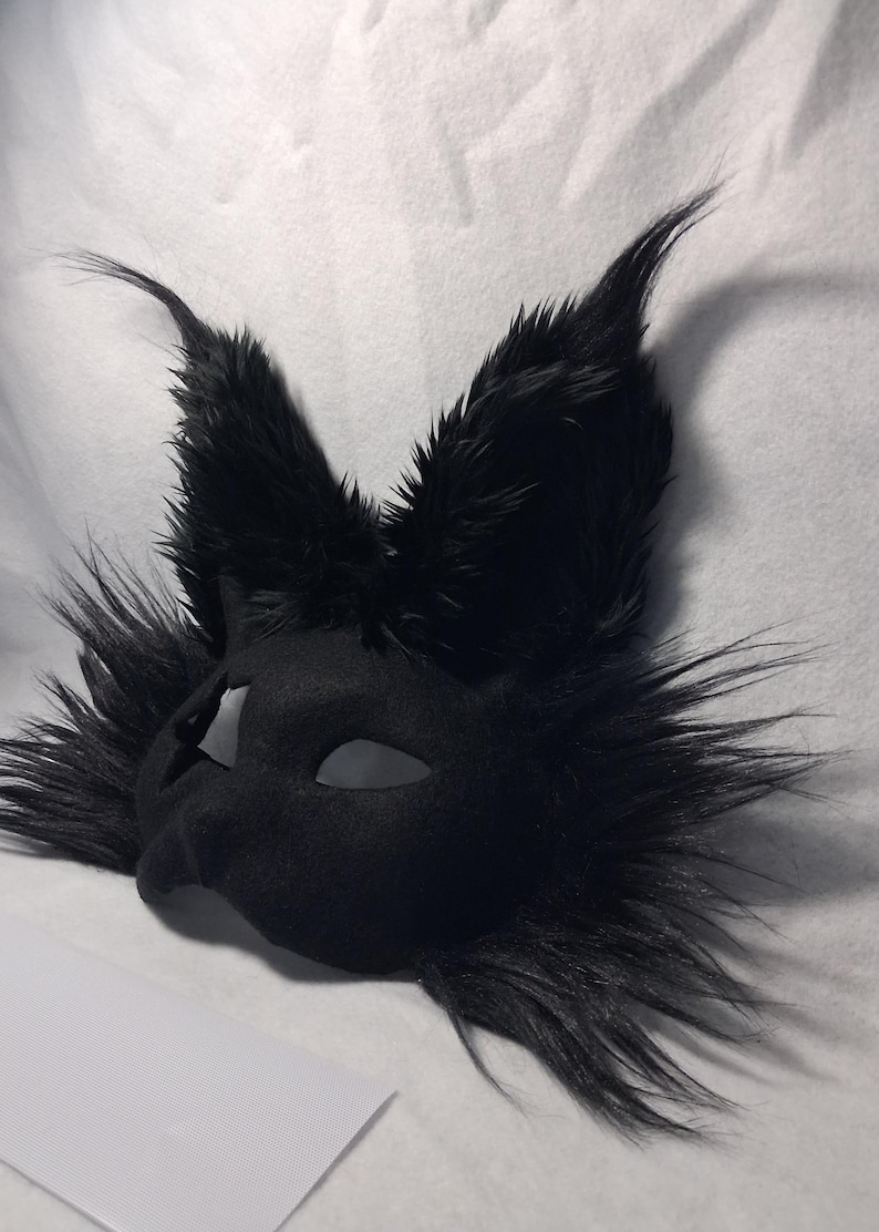 May include: A black, furry mask with pointed ears and eye cutouts. The mask has a smooth, matte finish and is surrounded by long, textured fur. The ears are upright and prominent, giving the mask a stylized animal appearance.