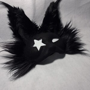 May include: A black mask with a star cutout and white eye holes. The mask is covered in black faux fur, with long, pointed ears. The mask is likely for a costume or cosplay.