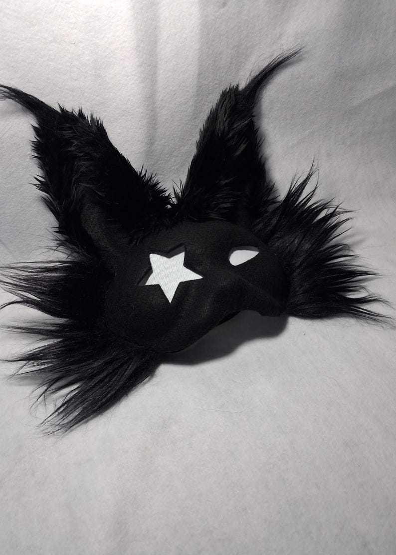 May include: A black mask with a star cutout and white eye holes. The mask is covered in black faux fur, with long, pointed ears. The mask is likely for a costume or cosplay.