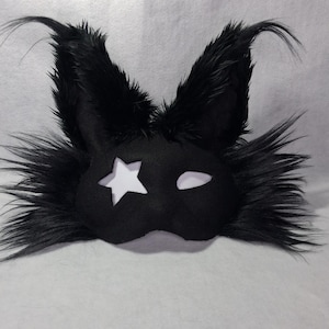 May include: Black eye mask with a star cutout and a white eye shape. The mask has long, fluffy black fur around the edges and on the ears. The mask is made of a soft material.