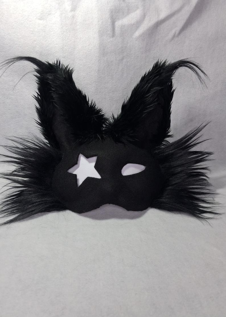 May include: Black eye mask with a star cutout and a white eye shape. The mask has long, fluffy black fur around the edges and on the ears. The mask is made of a soft material.