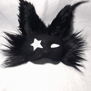 May include: Black felt mask with a star-shaped cutout on one eye and a curved slit for the other. The mask is trimmed with long, fluffy black faux fur, giving it a whimsical, animalistic appearance. The mask is designed for costume or cosplay.