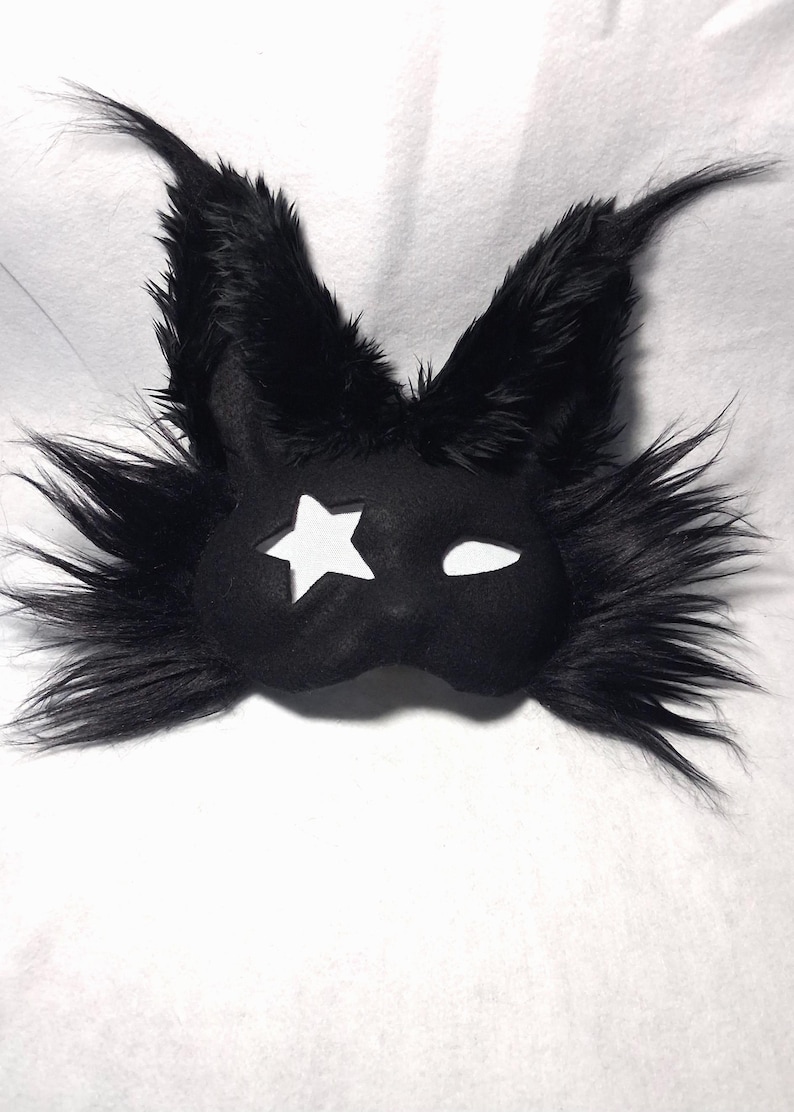 May include: Black felt mask with a star-shaped cutout on one eye and a curved slit for the other. The mask is trimmed with long, fluffy black faux fur, giving it a whimsical, animalistic appearance. The mask is designed for costume or cosplay.