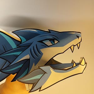 May include: A two-dimensional paper cutout of a shark-like creature head in profile. The design features shades of blue, white, and teal with sharp, pointed teeth, a dark blue eye, and distinct fin-like structures along the back of the head.