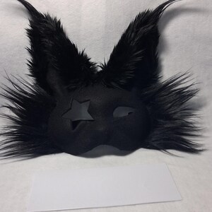 May include: Black furry mask with pointed ears and a star-shaped cutout on one eye. The mask has a sleek, dark design, perfect for costume parties or theatrical performances. The mask is made of a soft, plush material.