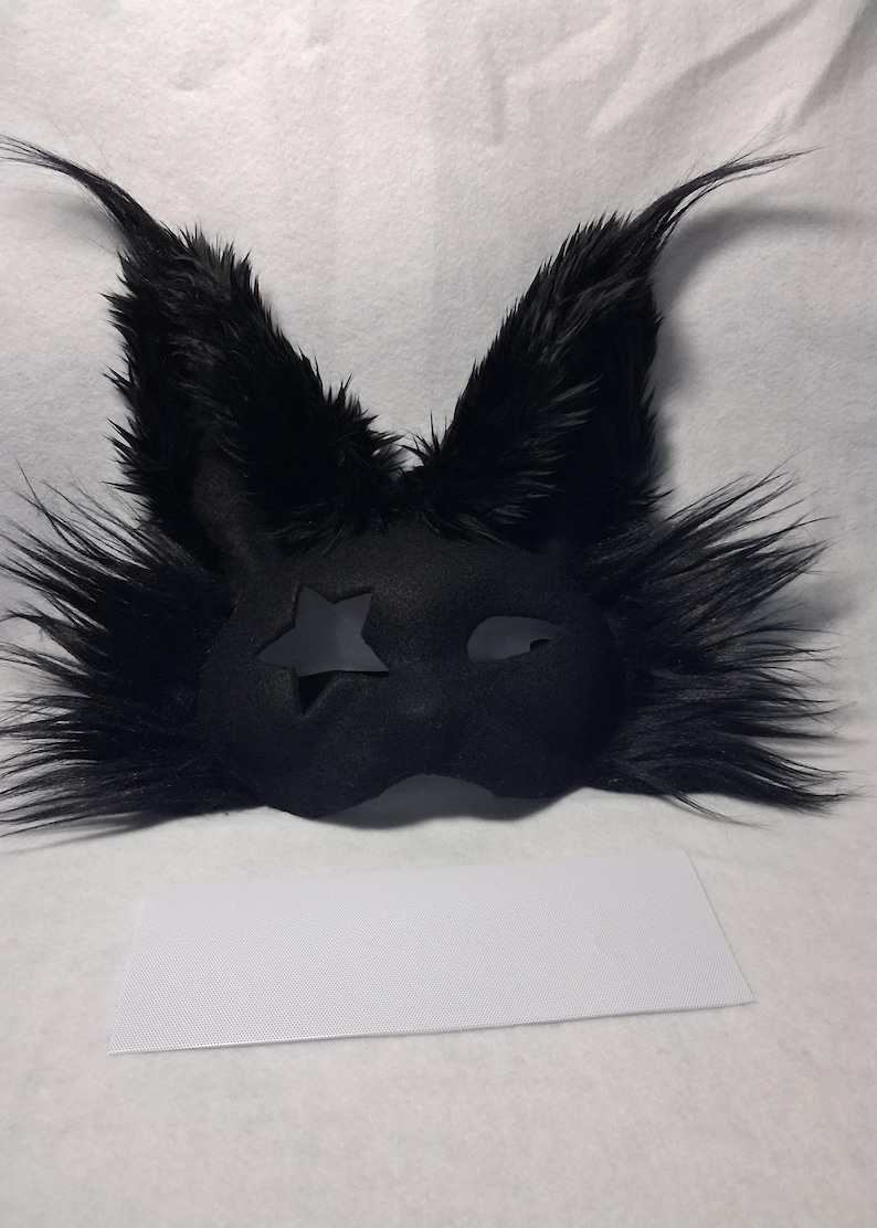 May include: Black furry mask with pointed ears and a star-shaped cutout on one eye. The mask has a sleek, dark design, perfect for costume parties or theatrical performances. The mask is made of a soft, plush material.