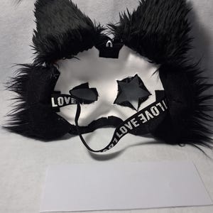 May include: A white mask with black accents, featuring star-shaped cutouts and fluffy black ears. The mask has a strap with the word "LOVE" printed on it. A white rectangular piece of fabric is below the mask.