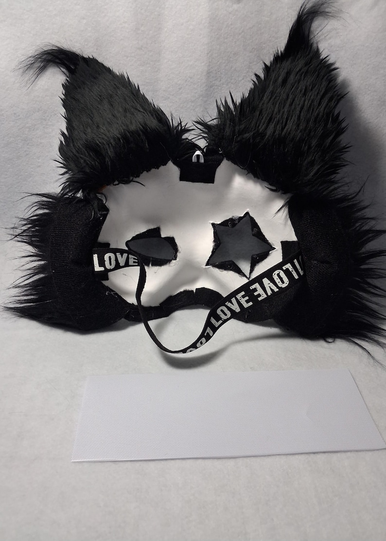 May include: A white mask with black accents, featuring star-shaped cutouts and fluffy black ears. The mask has a strap with the word "LOVE" printed on it. A white rectangular piece of fabric is below the mask.