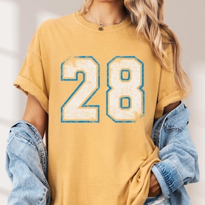 Puede incluir: Camiseta de color amarillo mostaza con el n&uacute;mero 28 en una fuente desgastada con contorno azul claro. La camiseta tiene mangas cortas y un corte relajado. Se lleva una chaqueta vaquera encima.