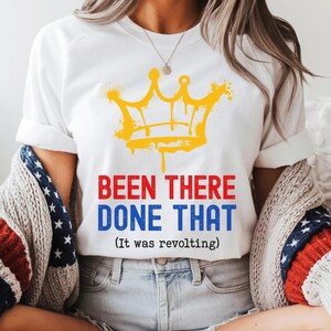May include: White t-shirt with a yellow crown graphic and the text "BEEN THERE DONE THAT (It was revolting)". The shirt is paired with denim shorts and a patriotic-themed sweater. Brown cowboy boots complete the look.