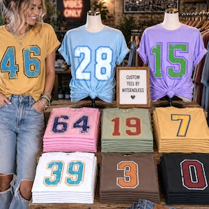 Puede incluir: Exhibici&oacute;n de camisetas personalizadas con n&uacute;meros, incluyendo una amarilla con el n&uacute;mero 46, una celeste con el 28 y una morada con el 15. Las camisetas est&aacute;n dobladas y apiladas en una exhibici&oacute;n de madera. El texto "GOOD VIBES ONLY" es visible.