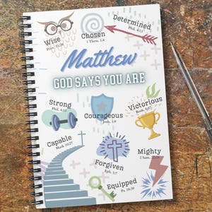 May include: A spiral-bound notebook with the title "Matthew" and the phrase "God Says You Are" in blue lettering. The notebook features various inspirational words and symbols, including "Wise," "Strong," and "Victorious," alongside related Bible verses. A pen rests beside the notebook.