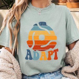 May include: A light blue t-shirt with a retro-style graphic of a record with the word "ADAPT" printed below it.