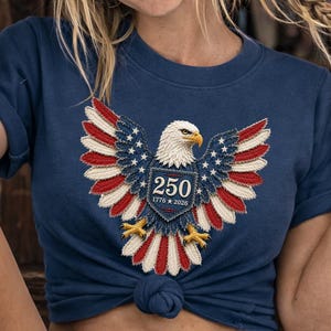 May include: A navy blue short-sleeved t-shirt featuring a central graphic of a bald eagle with wings spread, colored in red, white, and blue. The eagle holds a shield displaying the text 250 and the years 1776 and 2026 in white lettering.