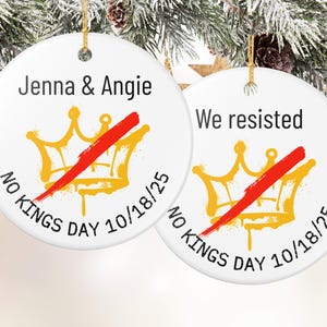 Puede incluir: Dos adornos redondos de cer&aacute;mica blanca con hilo dorado para colgar. Cada adorno presenta un dise&ntilde;o de corona dorada tachado con una raya roja. El texto incluye "Jenna & Angie" y "We resisted", con "No Kings Day 10/18/25".