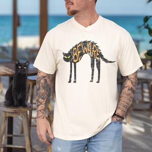 May include: Cream-colored t-shirt featuring a black cat design with the word "BEWARE" in orange. The cat has green eyes and a distressed, vintage look. A black cat sits on a wooden stool.