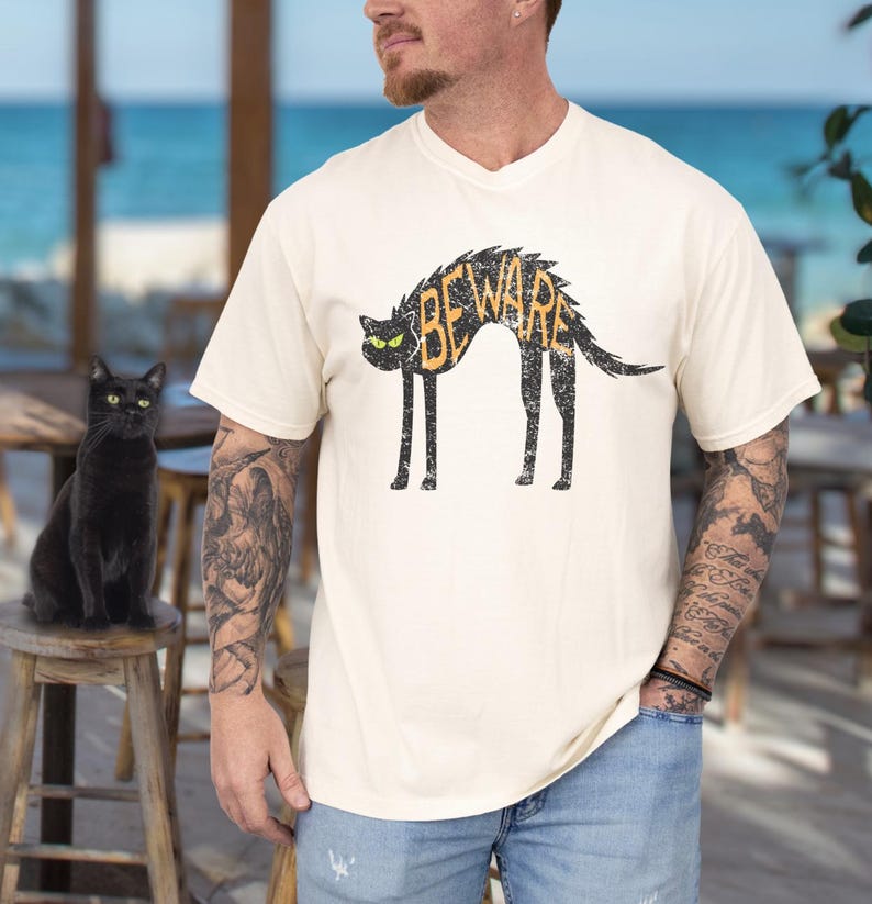 May include: Cream-colored t-shirt featuring a black cat design with the word "BEWARE" in orange. The cat has green eyes and a distressed, vintage look. A black cat sits on a wooden stool.