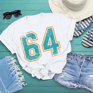 Puede incluir: Camiseta blanca con el n&uacute;mero "64" en turquesa y naranja, con un aspecto desgastado. Se combina con pantalones cortos vaqueros, gafas de sol, un sombrero para el sol y chanclas, evocando el verano.