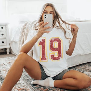 Puede incluir: Camiseta blanca con el n&uacute;mero 19 en un dise&ntilde;o rojo y amarillo desgastado. La persona que lleva la camiseta est&aacute; sentada en una alfombra estampada y se hace una foto con el tel&eacute;fono.
