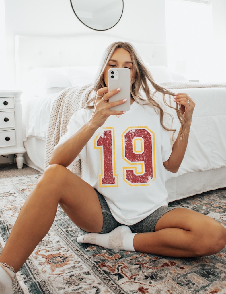 Puede incluir: Camiseta blanca con el n&uacute;mero 19 en un dise&ntilde;o rojo y amarillo desgastado. La persona que lleva la camiseta est&aacute; sentada en una alfombra estampada y se hace una foto con el tel&eacute;fono.