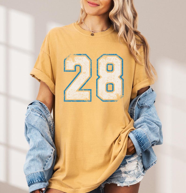 Puede incluir: Camiseta de color amarillo mostaza con el n&uacute;mero 28 en una fuente desgastada con contorno azul claro. La camiseta tiene mangas cortas y un corte relajado. Se lleva una chaqueta vaquera encima.