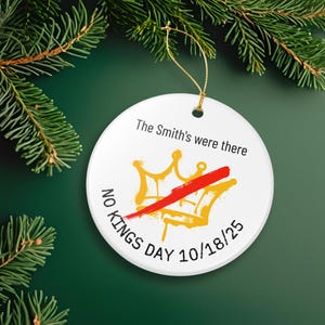 Puede incluir: Adorno navide&ntilde;o redondo de cer&aacute;mica blanca con una cuerda dorada, con un dise&ntilde;o de corona dorada tachada con una raya roja. El texto dice "The Smith's were there", "NO KINGS DAY 10/18/25". Sobre un fondo verde con ramas de pino.